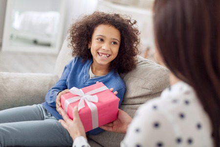 Beautiful joyful curly-haired girl sitting on the sofa and smiling and her mother giving her a giftの写真素材