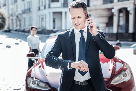 What time is it. Nice handsome pleasant businessman standing in front of his car and looking at the watch while talking on the phoneの写真素材