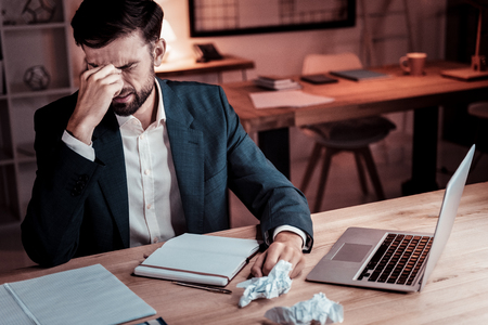 Upset disappointed unshaken worker sitting by the table opposite the laptop closing eyes and touching to his head.の写真素材