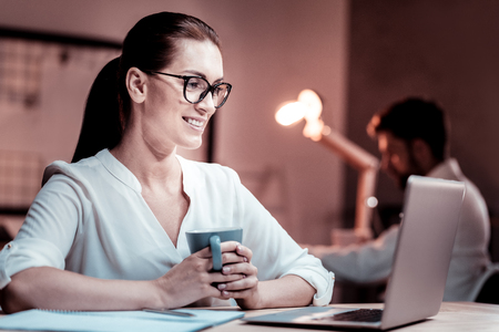 Good job. Satisfied bespectacled occupied woman sitting in the office by the table holding a cup and using the laptop.の写真素材