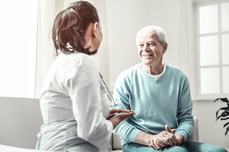 Appointment today. Senior kind nice-looking man sitting in the room at the sofa smiling and having consultation with a nurse.の写真素材