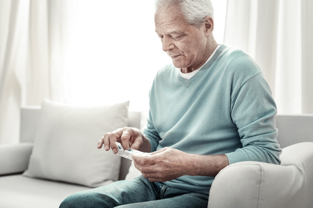 For healthy. Aged serious pleasant man sitting on the sofa taking a pill and looking at tablets.の写真素材