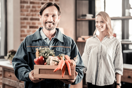 Cheerful nice handsome man holing a box with vegetables and smiling while delivering itの写真素材