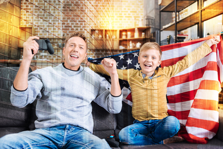 Enthusiastic players. Emotional active young man putting his hands up and shouting while his cheerful smiling son sitting by his side with a big American flag and celebrating their victory in a gameの写真素材