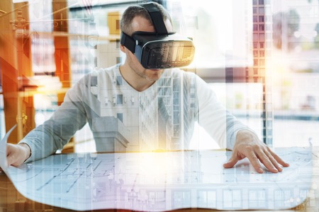New drawing. Clever experienced architect sitting at the table and wearing virtual reality glasses while working with a large detailed drawing being placed in front of himの写真素材