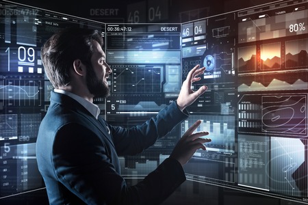 Easy work. Clever experienced professional programmer smiling and feeling glad while standing in front of a transparent screen and controlling the programの写真素材