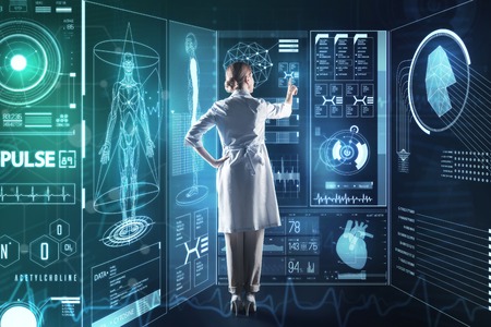 Progressive medicine. Clever qualified medical worker looking concentrated while standing in front of a giant transparent screen and using futuristic technologies in her workの写真素材