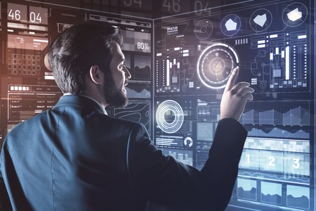 Interesting information. Young cheerful bearded programmer looking interested while touching a transparent screen and reading the information on itの写真素材
