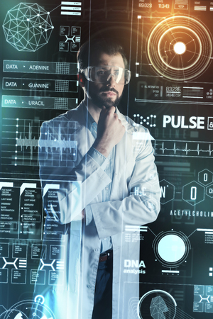 Interesting idea. Calm smart medical worker standing with his fingers touching the beard and thoughtfully looking at the information on a transparent screenの写真素材
