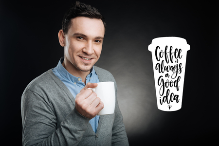 Amazing coffee. Calm handsome young man having a productive working day and feeling glad while having a short rest and drinking hot tasty coffee from his big mugの写真素材