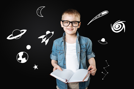Curious boy. Clever cheerful curious boy wearing big glasses and feeling glad while holding a big notebook and reading the information about the spaceの写真素材