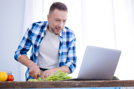 Anti aging nutrition. Focused serious appealing man gazing at screen while chopping salad and using knifeの写真素材