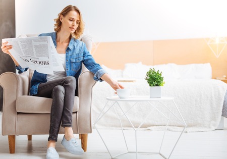 Favorite activity. Calm thoughtful beautiful woman sitting in a soft armchair and holding a big interesting newspaper while trying to take a cup of tea from the little table next to herの写真素材