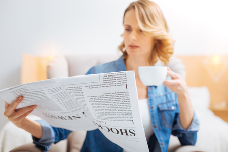Looking worried. Young smart attentive woman sitting in her bedroom with a cup of tasty hot tea and feeling worried while reading the latest news in a local newspaperの写真素材