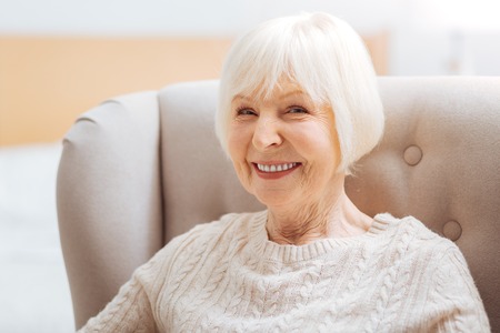 Pretty pensioner. Cute pretty senior woman feeling happy while sitting in a comfortable soft armchair and waiting for her kind young granddaughter to come and visit herの写真素材
