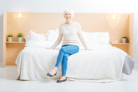 Happiness. Pleasant kind cheerful aged woman smiling and looking happy while being in a beautiful light comfortable room and sitting on a nice soft bedの写真素材
