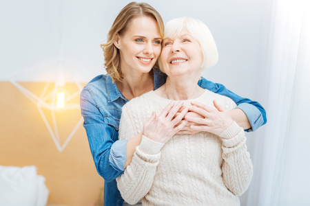 Feeling warmth. Beautiful young kind woman enjoying spending time with her positive cheerful grandmother while standing and hugging herの写真素材