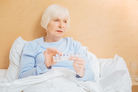 Useful box. Calm serious aged woman feeling bad and looking into the distance while staying in bed with a convenient pillbox in her handsの写真素材