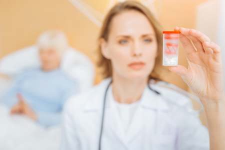 New pills. Smart experienced professional doctor looking at the box with new pills while her ill tired patient staying in bed behind her backの写真素材
