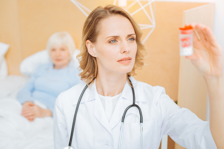 Careful doctor. Skilled professional experienced doctor feeling confident while standing next to her aged ill patient and looking at the little box with necessary pillsの写真素材