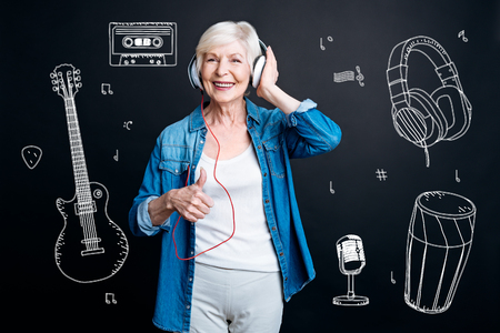 Music lover. Emotional positive aged lady feeling happy and smiling while listening to loud music in her convenient headphones and cheerfully putting her thumb upの写真素材