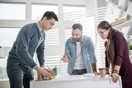 Thinking together. Attractive concentrated young bearded man holding a pencil and standing at the table with his colleagues and discussing work with themの写真素材