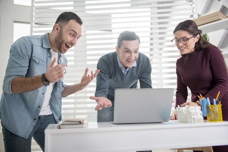 Wonderful news. Good-looking alert young dark-haired man smiling and pointing at the laptop and his...