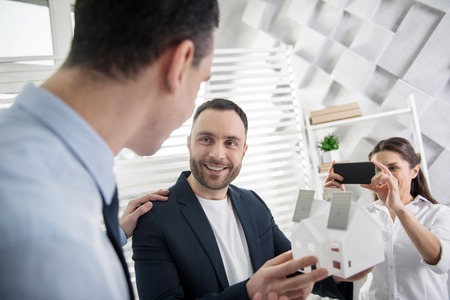 I feel happy. Attractive content young dark-haired architect smiling and holding a house miniature while their colleague taking photos of themの写真素材