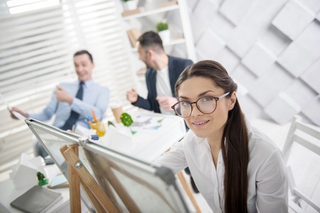 Good day. Beautiful alert young long-haired woman wearing glasses and working on the project while her colleagues talking in the backgroundの写真素材