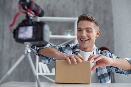 I feel happy. Handsome cheerful young well-built man smiling and opening his box while making a videoの写真素材