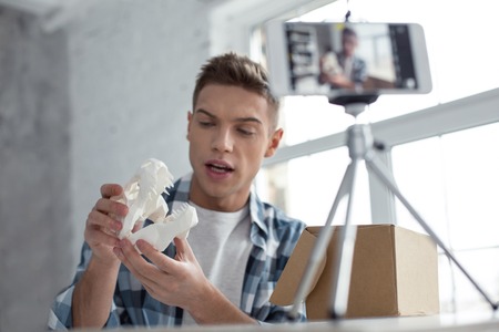 Interesting theme. Handsome concentrated young well-built man talking and showing a dinosaur skull while making a videoの写真素材