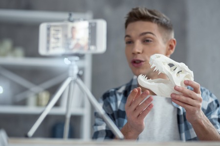 My hobby. Attractive joyful young well-built man talking and showing a dinosaur skull while making a videoの写真素材