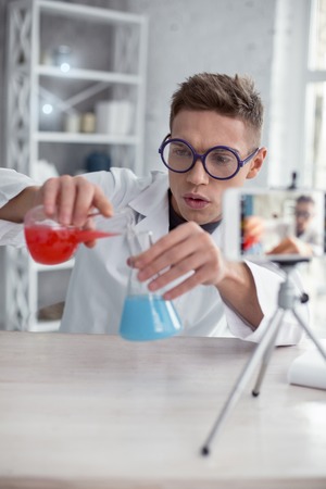 Expecting results. Handsome inspired fair-haired teenager wearing a uniform and glasses and holding test tubes while making a videoの写真素材