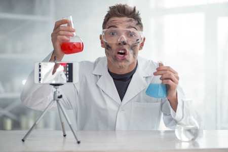 It is amazing. Handsome impressed dirty fair-haired young blogger and wearing uniform and holding test tubes while making new videoの写真素材