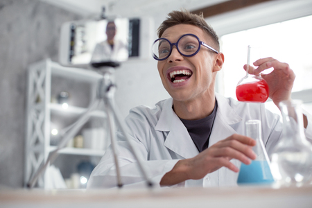 In high spirits. Attractive exuberant fair-haired teenager wearing a uniform and glasses and holding test tubes while doing an experimentの写真素材