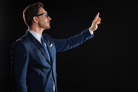 Important detail. Occupied beardful stylish man standing in the dark room looking aside and touching to the invisible object.の写真素材