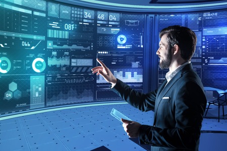 Convenient innovation. Cheerful calm young programmer smiling and feeling satisfied with his convenient gadgets while standing in front of a futuristic computer with a transparent tablet in his handの写真素材