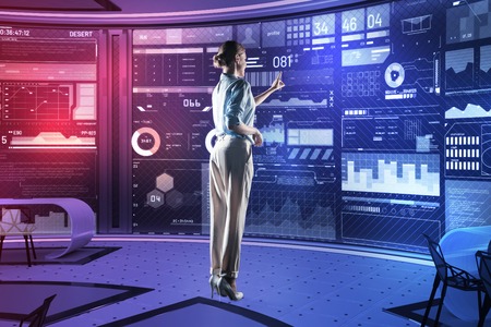 Young specialist. Smart experienced professional programmer standing in front of a transparent futuristic device and pointing to the screen of it while being in her equipped officeの写真素材