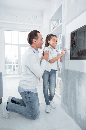 Do not worry. Nice positive handsome man standing near his daughter and helping her while teaching how to use smart home technologyの写真素材