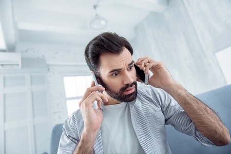 It is not truth. Surprised bearded male person wrinkling forehead while holding telephones near ears...