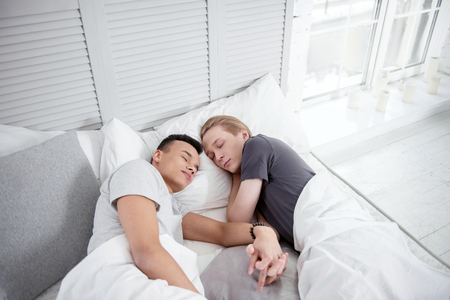 Always together. Pretty sweet gay couple sleeping while holding hands and lying on bedの写真素材