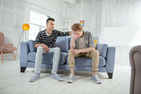 Problem in life. Sad depressed gay couple sitting on sofa while man calming down guy who cryingの写真素材