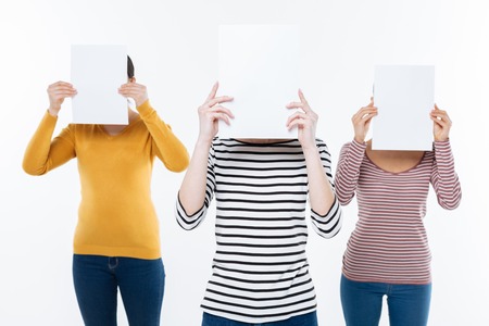 You dont see us. Nice delighted young women standing against white background and holding sheets of paper while hiding their facesの写真素材