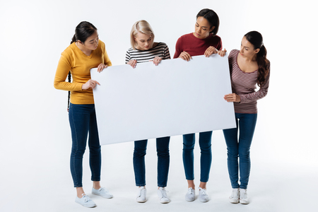 Our slogan. Smart joyful confident women standing around the poster and looking at it while holding it in their handsの写真素材