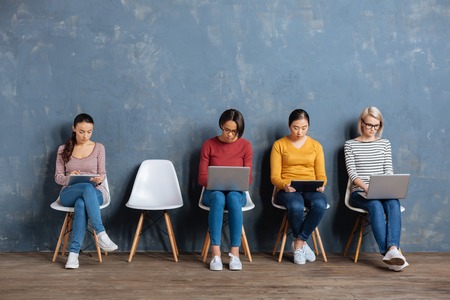 Modern generation. Nice pleasant young women sitting on the chairs and using their devices while surfing the Internetの写真素材