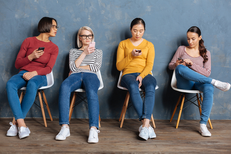Digital gadgets. Attractive nice pretty women holding their smartphones and using them while sitting on the chairsの写真素材