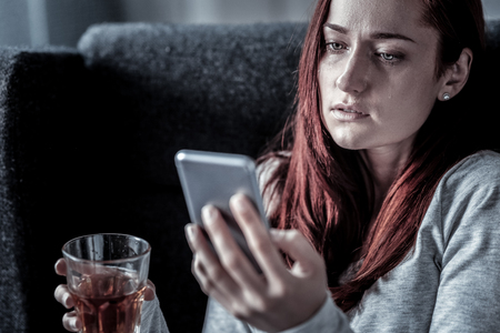 Leave me alone. Stressful upset redhead woman sitting on the chair drinking and using cellphone.の写真素材