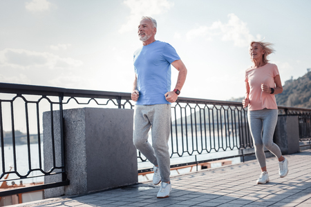 Healthy is the most important. Joyful aged confident man running across the quay with a woman spending time on fresh air and doing cardio exercises.の写真素材