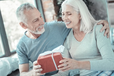 Im so happy. Satisfied gray haired pretty woman sitting in the room near her husband taking the present and rejoicing.の写真素材