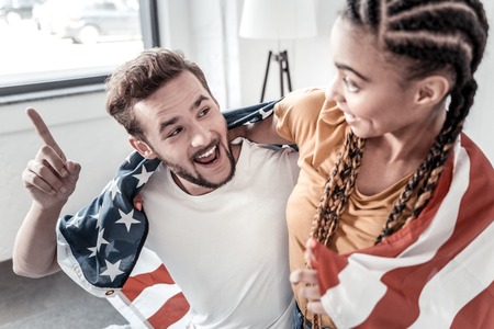Patriotic couple. Joyful positive young couple sitting together and being wrapped in the US flag while expressing their patriotic feelingsの写真素材